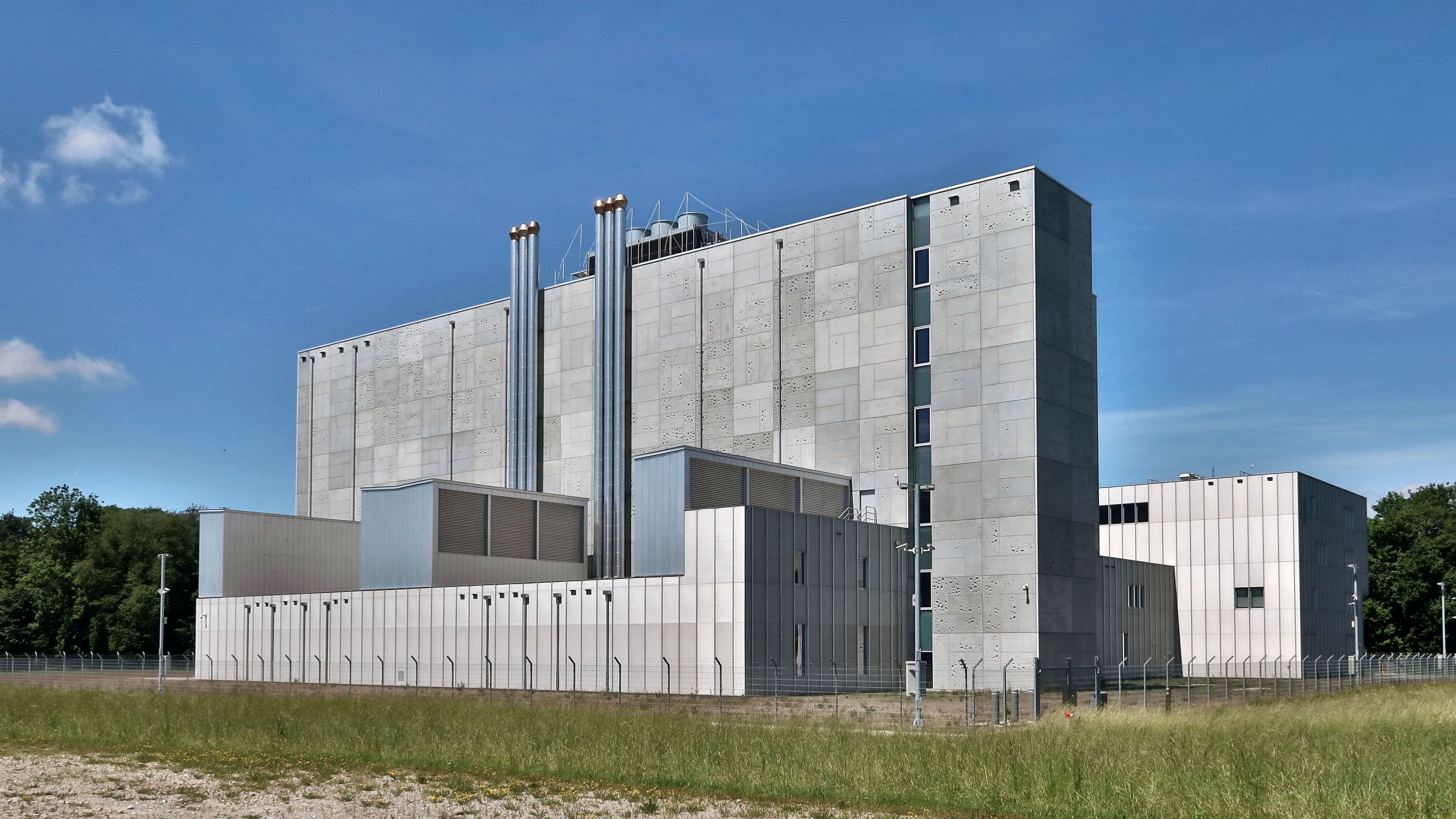 Grey concrete data centre building with tall exhaust stacks and a perimeter fence, beside a grass field under a blue sky in Frauenfeld, Switzerland