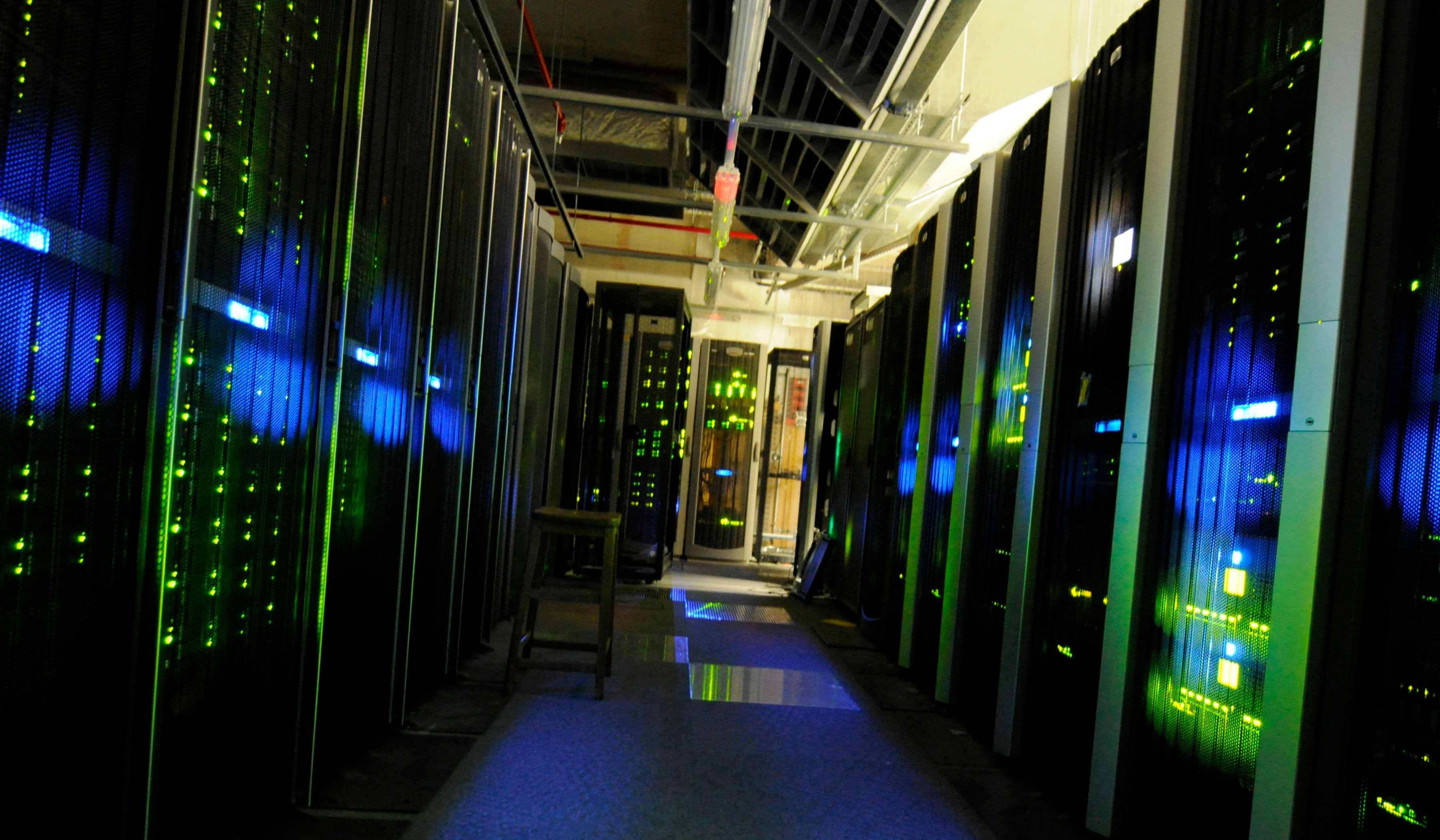 A narrow aisle between rows of dark server racks with blue and green status lights and overhead cable trays