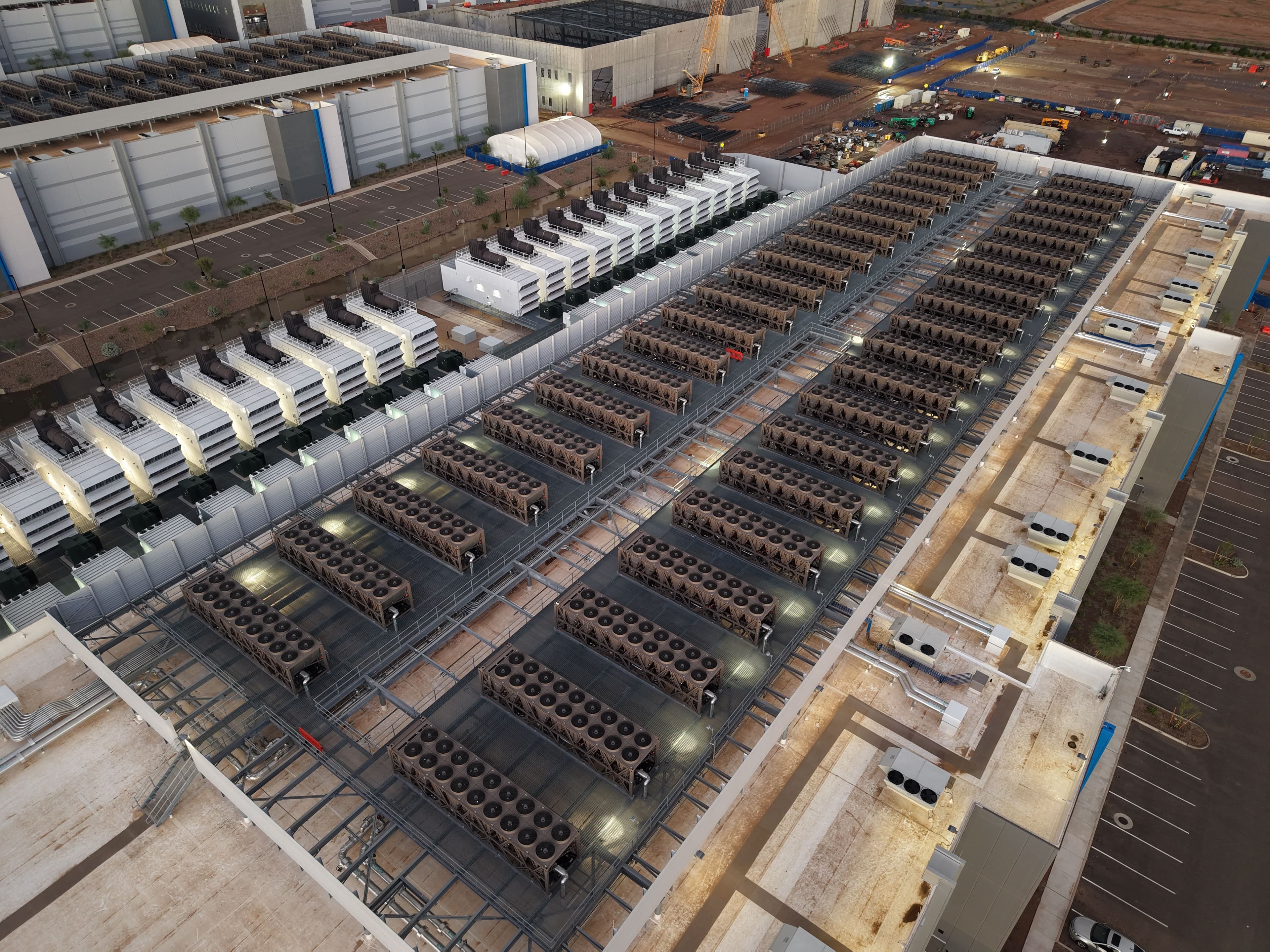 Aerial view of a data centre roof at dusk with rows of cooling units and backup generators, beside a car park and buildings under construction